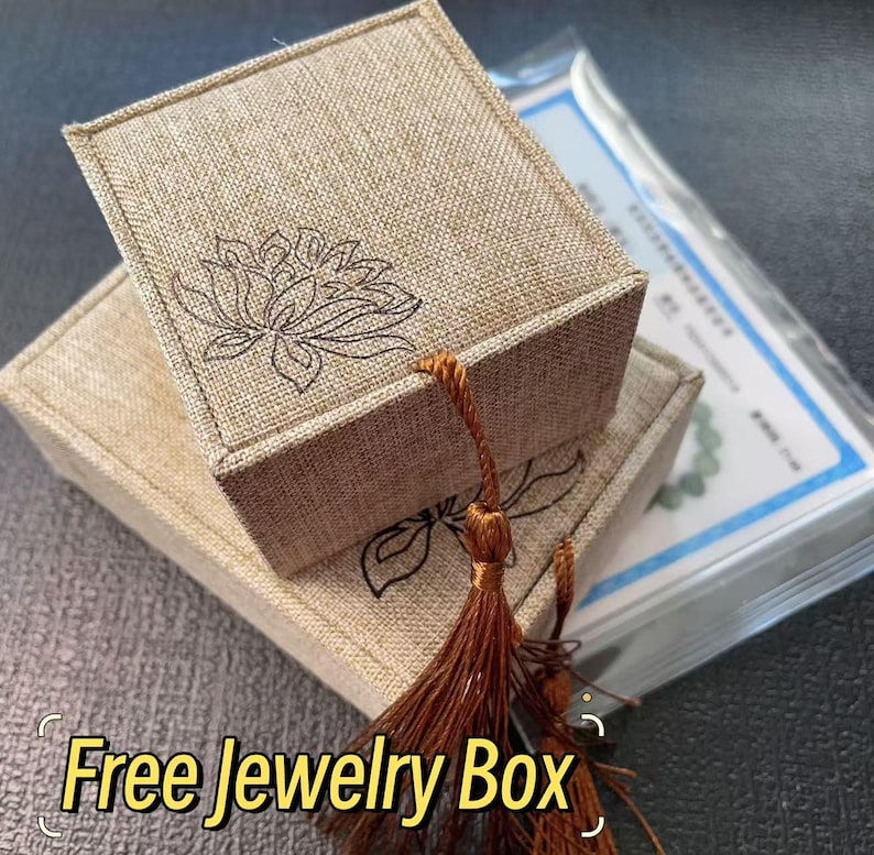 May include: A square, tan, textured fabric jewelry box with a lid featuring a black line-art lotus flower design. A brown silk tassel hangs from the corner of the lid. The box sits atop a matching rectangular base, with a clear plastic package nearby.