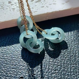 Peut inclure: Un collier délicat avec plusieurs anneaux de jade bleu clair reliés entre eux. Les anneaux sont suspendus à une chaîne dorée, créant un effet superposé. Le jade a une qualité translucide. Le collier est présenté sur une surface texturée.