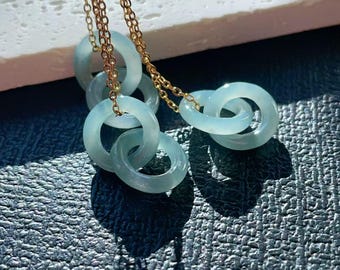 May include: A necklace with multiple light blue jade rings linked together. The rings are suspended from a gold chain, creating a layered effect. The jade has a translucent quality. The necklace is displayed on a textured surface.