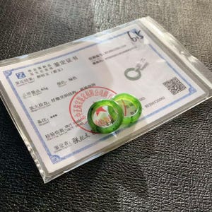 May include: Two translucent green jade rings are displayed on a certificate. The rings have a circular design with a polished finish. The certificate includes Chinese text and a red stamp, along with a clear plastic bag.