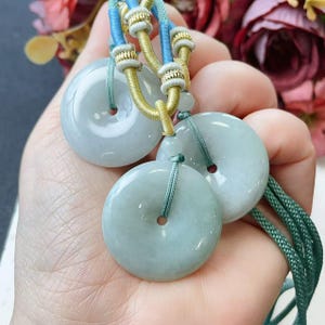 May include: Three pale green jade donut pendants with blue, gold, and green cords. Each pendant has a central hole. The pendants are held by a hand. The background includes colorful flowers.