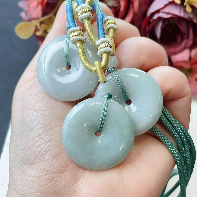 May include: Three pale green jade donut pendants with blue, gold, and green cords. Each pendant has a central hole. The pendants are held by a hand. The background includes colorful flowers.