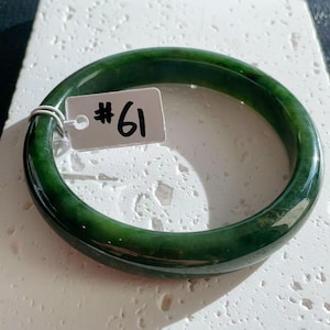 May include: A dark green jade bangle. The polished stone is a deep green colour. A small tag with the number "61" is attached. The bangle is circular in shape.