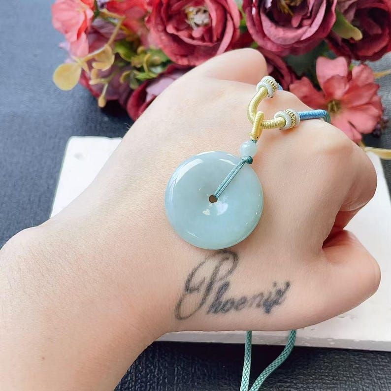 May include: A circular, pale green jade pendant with a central hole, suspended by a teal cord and a decorative knot. The pendant is smooth and polished, with a subtle translucence. The cord is attached to a yellow and blue decorative knot.
