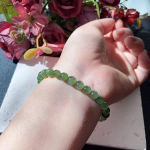 May include: A green jade bead bracelet. The bracelet features round, polished beads. The beads are a translucent green colour. The bracelet is worn on a wrist. The background includes flowers.