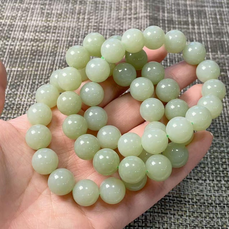 May include: A stretch bracelet composed of numerous smooth, round, pale green beads. The beads have a polished, semi-translucent finish and are strung together in a circular formation, creating a continuous loop of light green stone spheres.