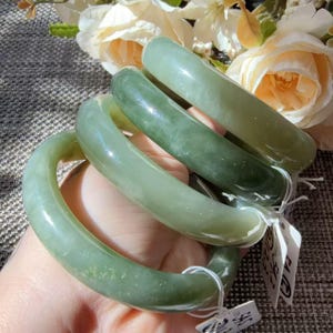 May include: Three smooth, polished jade-colored bangle bracelets with a rounded profile. Each bracelet features a translucent, light green hue with subtle variations in tone and texture. Small white paper tags are attached to the bangles.