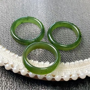 May include: Three polished, green jade rings. The rings are a vibrant shade of green with a smooth, glossy finish. The rings are displayed on a white lace surface with a pearl trim.