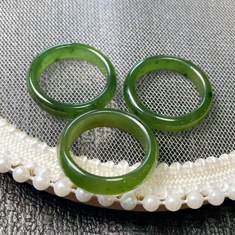 May include: Three polished, green jade rings. The rings are a vibrant shade of green with a smooth, glossy finish. The rings are displayed on a white lace surface with a pearl trim.