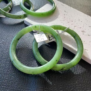 Könnte beinhalten: Zwei grüne Jade-Armreifen mit einem gesprenkelten Muster. Die Armbänder sind kreisförmig und haben eine glatte, polierte Oberfläche. An jedem Armreif ist ein Preisschild befestigt. Der Hintergrund ist eine Mischung aus schwarzen und weißen Oberflächen.