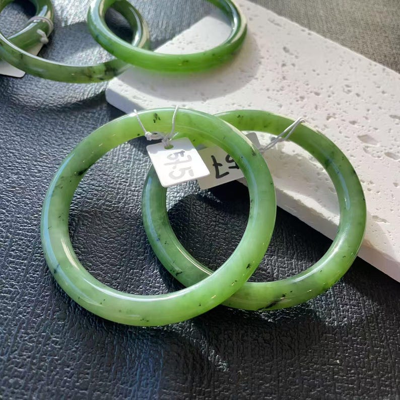 May include: Two green jade bangles with a speckled pattern. The bracelets are circular and have a smooth, polished finish. Each bangle has a price tag attached. The background is a mix of black and white surfaces.