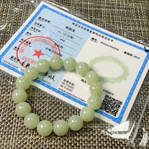 May include: A pale green jade bead bracelet consisting of smooth, round, polished stones arranged in a circular loop. The bracelet is displayed on top of a white rectangular certificate of authenticity featuring black text and a red star stamp.
