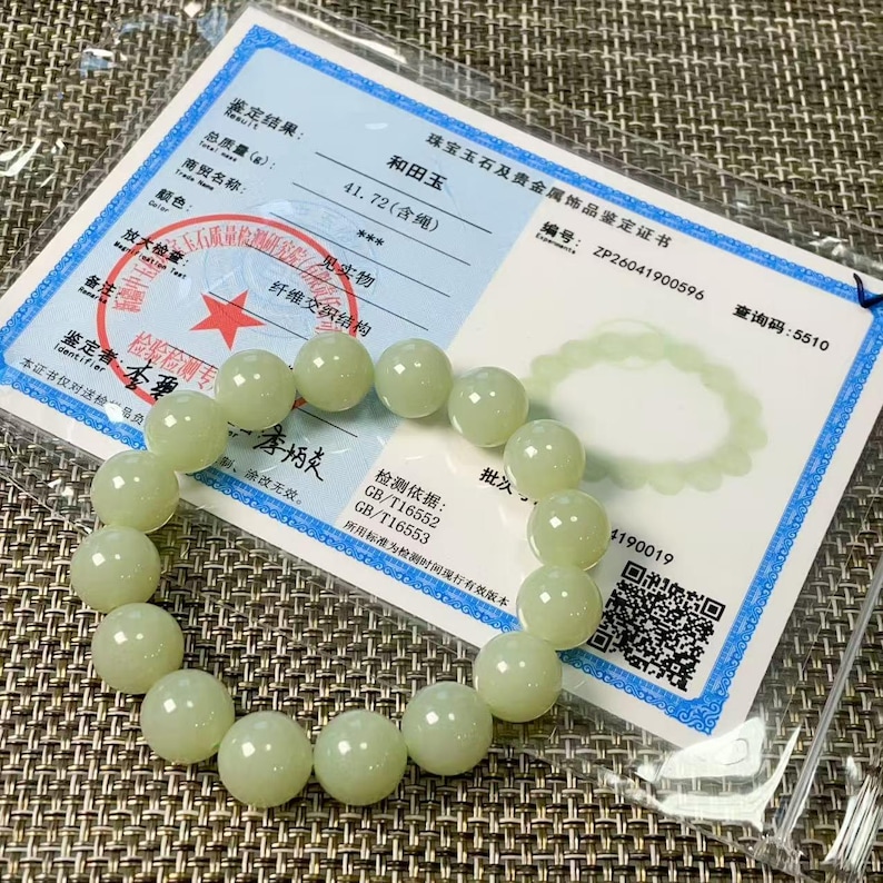 May include: A pale green jade bead bracelet consisting of smooth, round, polished stones arranged in a circular loop. The bracelet is displayed on top of a white rectangular certificate of authenticity featuring black text and a red star stamp.