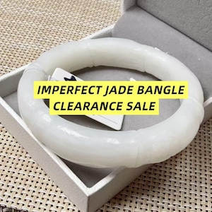Pode incluir: Um bracelete de jade branco com uma textura lisa e segmentada que lembra bambu. Uma etiqueta branca está presa ao bracelete com o texto preto IMPERFECT JADE BANGLE CLEARANCE SALE impresso em um fundo amarelo sobre a peça de joalheria.