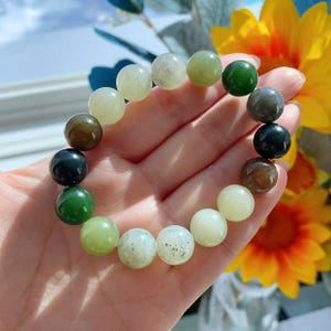 May include: A beaded bracelet featuring a variety of colorful, round gemstones. The bracelet includes beads in shades of green, brown, black, and cream. The bracelet is held in a hand, with a yellow flower in the background.