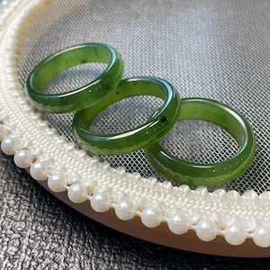 May include: Three polished green jade rings. The rings are a vibrant green color and appear smooth and glossy. They are displayed on a white lace-trimmed surface with small, round pearls.
