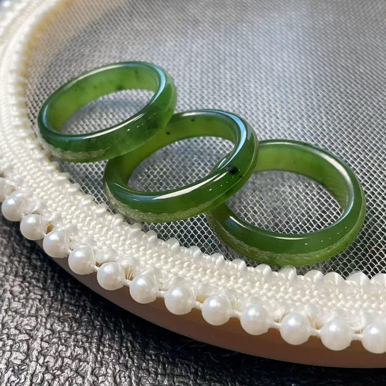 May include: Three polished green jade rings. The rings are a vibrant green color and appear smooth and glossy. They are displayed on a white lace-trimmed surface with small, round pearls.
