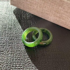 May include: Two green jade rings. The rings are a vibrant green color with darker green speckles. The rings are set against a textured black surface, with a neutral-toned box in the background.