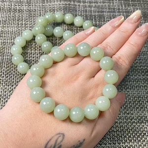 May include: A stretch bracelet composed of smooth, round, pale mint green beads. The beads have a polished, semi-translucent finish and are strung together in a circular loop. The bracelet is shown in a neutral, light-toned color palette.