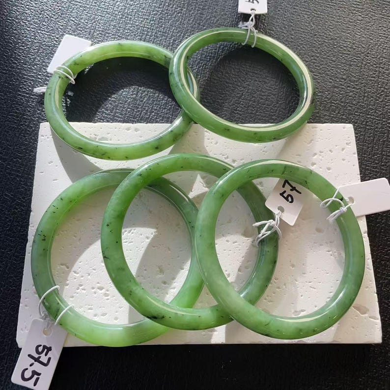 May include: Five jade bangles in varying shades of green, with dark speckles, are displayed on a white surface. The circular bracelets are smooth and polished, showcasing the natural variations in the stone. Each bangle is a complete circle, ready to be worn.