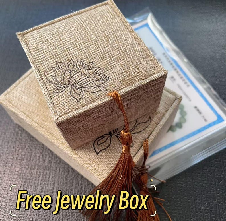May include: Two beige linen jewelry boxes stacked, one atop the other. Each box features a floral design and a decorative tassel. The text "Free Jewelry Box" is displayed in a yellow font at the bottom of the image.
