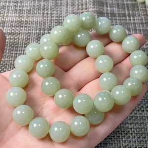 May include: A circular bracelet composed of smooth, polished, pale green jade beads strung together. The beads are uniform in size and shape, featuring a soft, translucent luster that reflects light across their rounded surfaces in a continuous loop.
