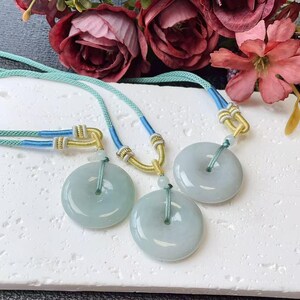 May include: Three jade donut pendants with light blue cords and gold accents. The jade is a pale green colour. The cords are light blue and green. The background includes flowers.