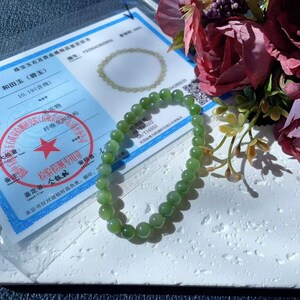May include: A light green jade bead bracelet. The bracelet is made of smooth, spherical beads. The image also shows a certificate with Chinese text and a floral arrangement with dark red roses.