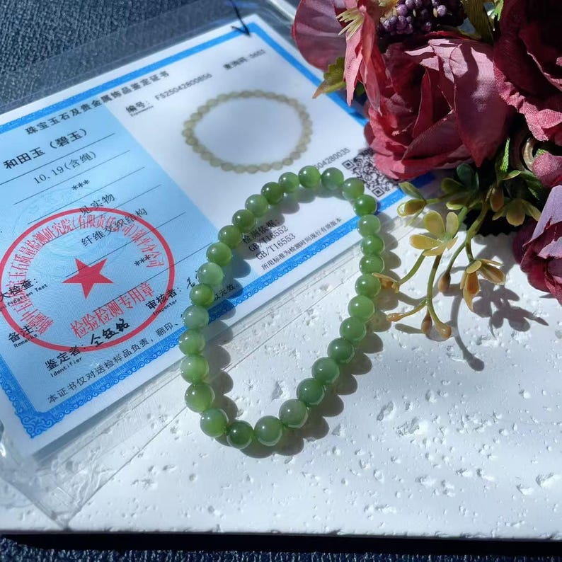 May include: A light green jade bead bracelet. The bracelet is made of smooth, spherical beads. The image also shows a certificate with Chinese text and a floral arrangement with dark red roses.