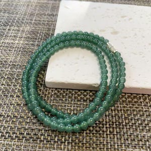 May include: A multi-strand necklace composed of small, smooth, translucent green beads. The beads are uniform in size and shape, arranged in a circular pattern. A small, cylindrical silver-toned metal clasp connects the ends of the beaded strands.