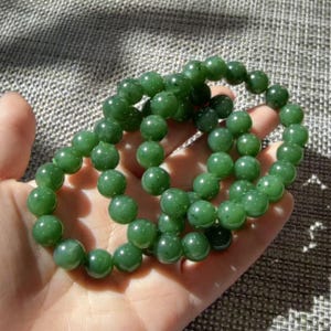 May include: A beaded bracelet composed of numerous smooth, spherical green jade beads strung together on an elastic cord. The beads exhibit varying shades of deep and forest green with subtle natural dark inclusions throughout the polished surface.