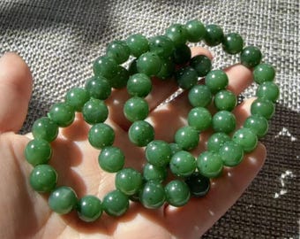 May include: A beaded bracelet composed of numerous smooth, spherical green jade beads strung together on an elastic cord. The beads exhibit varying shades of deep and forest green with subtle natural dark inclusions throughout the polished surface.