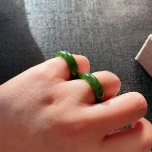 May include: Two green jade rings are worn on a hand. The rings are a vibrant, solid green color and appear to be smooth and polished. The rings are simple bands, showcasing the natural beauty of the jade.