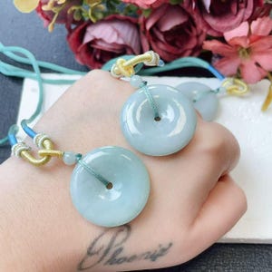 May include: Two pale green jade donut pendants with a central hole, strung on teal and gold cords. The pendants are smooth and polished, reflecting light. The background features colorful flowers.