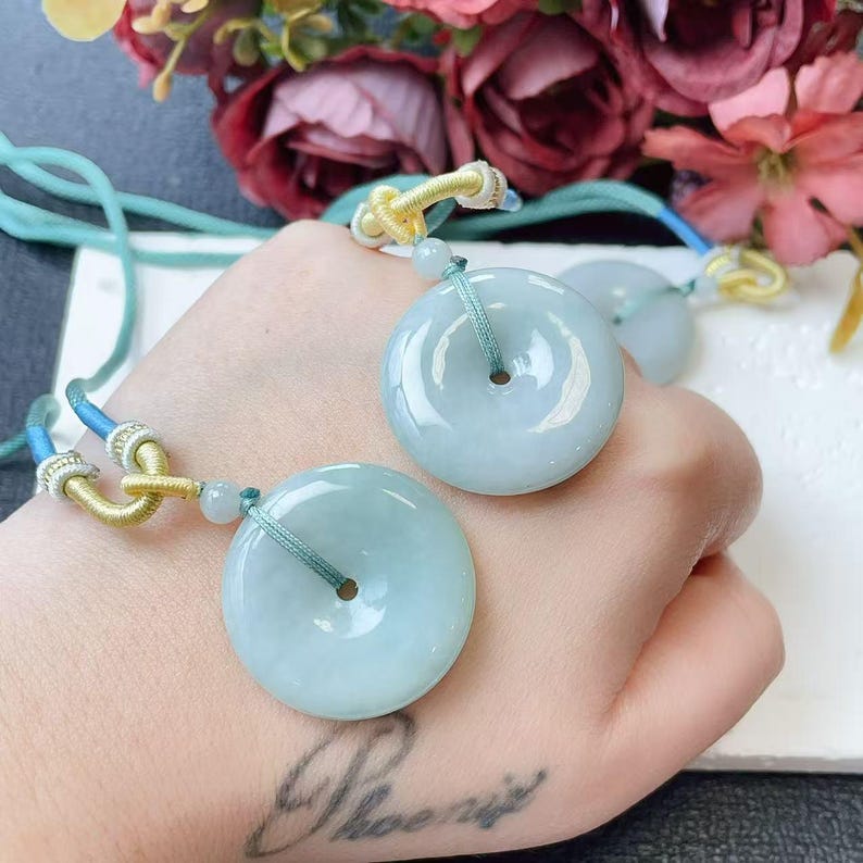 May include: Two pale green jade donut pendants with a central hole, strung on teal and gold cords. The pendants are smooth and polished, reflecting light. The background features colorful flowers.