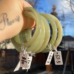 May include: Four pale green jade bangles are held in a hand. The smooth, translucent bracelets have varying shades of green. Price tags are attached to the bangles. The background is blurred, with a glimpse of a house and sky.