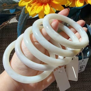 May include: A stack of five smooth, translucent white circular bangles. Each bangle has a rounded profile and a polished finish. Two small white rectangular tags are attached to the bangles, with one tag displaying the handwritten number 60.4.