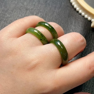 May include: Four jade rings in varying shades of green are displayed on fingers. The rings have a smooth, polished surface and a rounded shape. The jade appears to be translucent, with some rings showing small dark inclusions.
