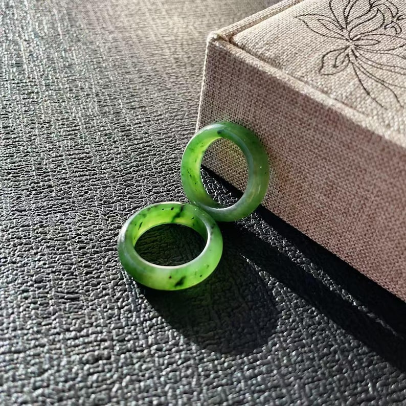 May include: Two green jade rings with varying shades of green and black speckles. The rings are circular and sit on a textured, dark surface next to a beige box with a floral design.