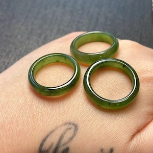 May include: Three green jade rings. The rings are a solid, smooth band of translucent green stone. The rings are displayed on a light skin tone background.