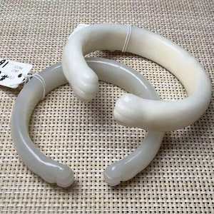May include: Two off-white, smooth, circular bracelets featuring carved animal paw motifs at the open ends. The bracelets have a polished, semi-translucent appearance and are secured with thin white plastic ties, with a small white price tag attached.