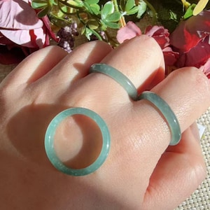 May include: Three smooth, polished jade rings in a pale, translucent green hue. Two of the rings are worn on fingers, while the third ring is positioned separately in the foreground, showcasing a simple, circular band design.