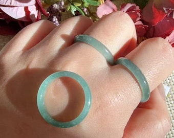 May include: Three smooth, polished jade rings in a pale, translucent green hue. Two of the rings are worn on fingers, while the third ring is positioned separately in the foreground, showcasing a simple, circular band design.