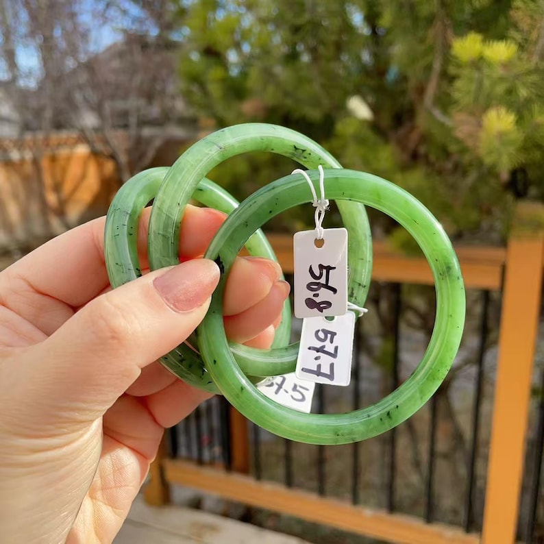 May include: Three jade green bangles with a translucent appearance. The bangles are round and smooth, with a slightly mottled texture. Each bangle has a tag attached with the numbers "56.8" and "57.7" printed on it.
