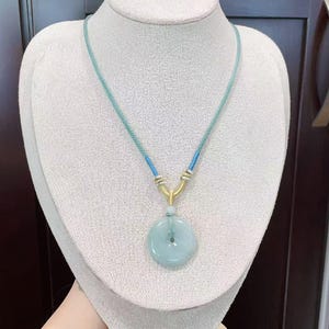 May include: A light blue jade pendant necklace. The pendant is a circular disc with a central hole, suspended from a blue and gold cord. The necklace is displayed on a white mannequin bust.