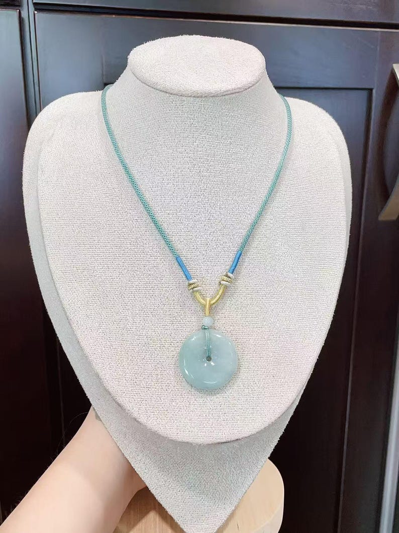 May include: A light blue jade pendant necklace. The pendant is a circular disc with a central hole, suspended from a blue and gold cord. The necklace is displayed on a white mannequin bust.