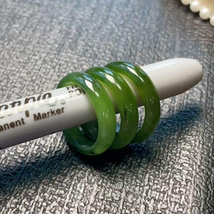May include: Three translucent green jade rings are displayed on a white marker. The rings have a smooth, polished finish and a vibrant green color. The marker has the word "Permanent" printed on it.