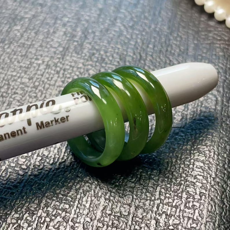 May include: Three translucent green jade rings are displayed on a white marker. The rings have a smooth, polished finish and a vibrant green color. The marker has the word "Permanent" printed on it.