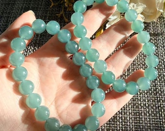 May include: A beaded bracelet featuring smooth, round, translucent light green gemstone beads strung on an elastic cord. The beads have a polished finish and a consistent pale mint green hue, creating a circular loop that rests against a palm.