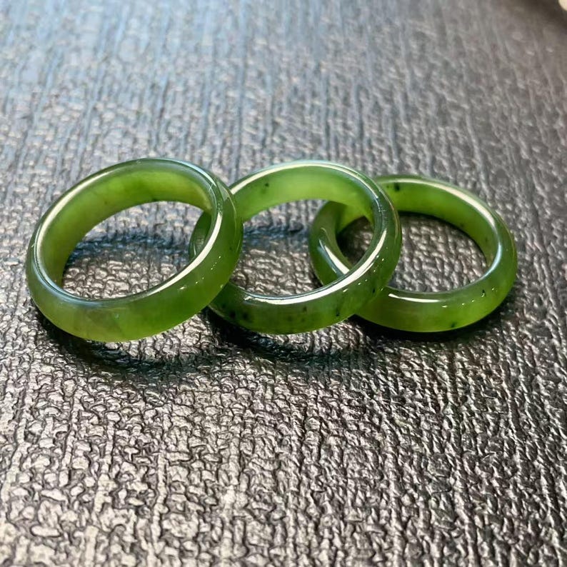 May include: Three green jade rings are displayed. The rings are a vibrant green color with subtle variations in tone and small dark inclusions. The rings are smooth and polished, reflecting light.
